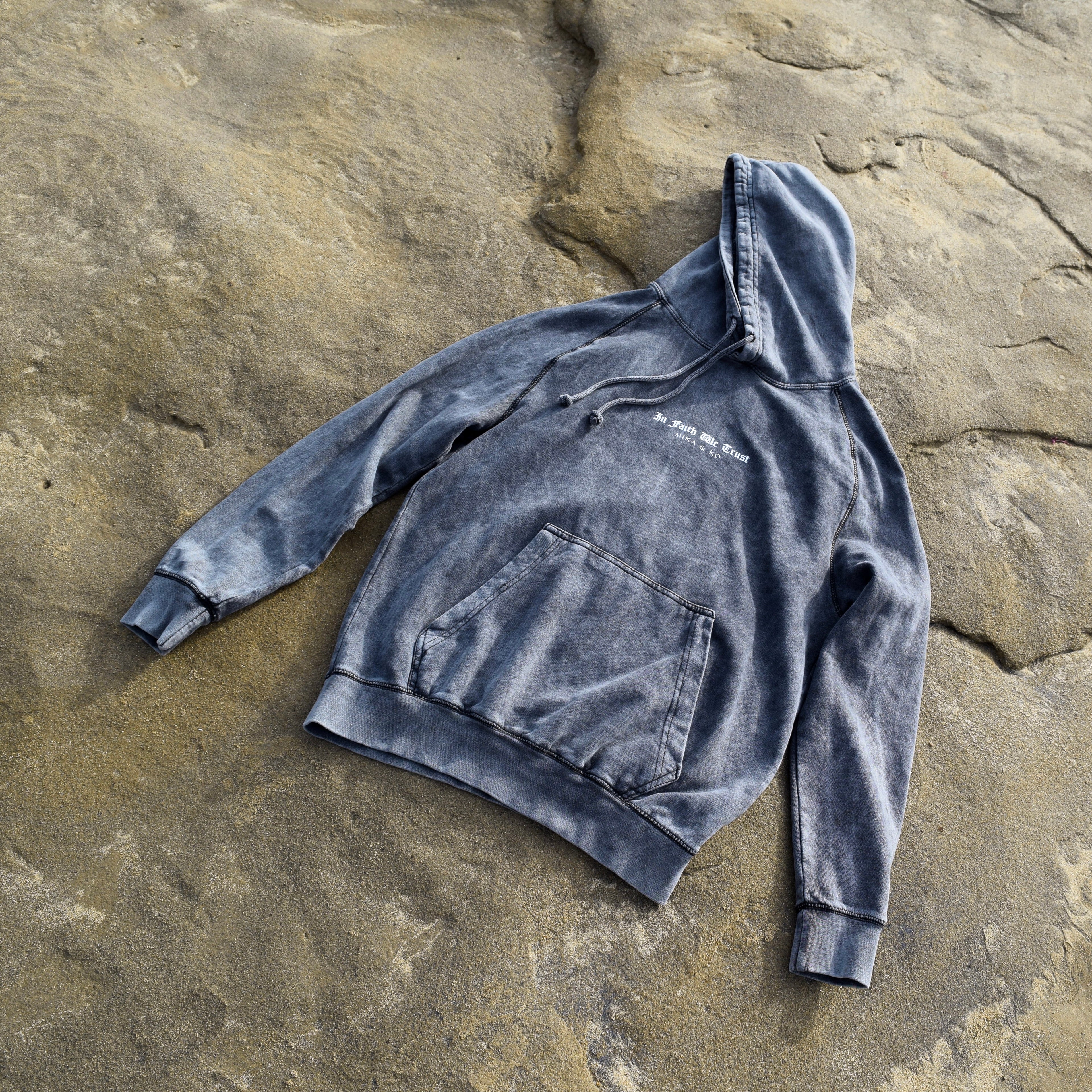 Front of In Faith We Trust Hoodie laying flat on a beach rock.